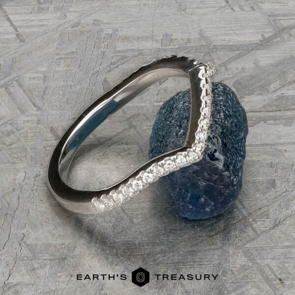 The "Graciella" band in 14k white gold The "Graciella" band in 14k white gold, featuring a curved, textured design, is displayed leaning against a rough blue gemstone on a patterned surface. The logo "Earth's Treasury" appears at the bottom.
