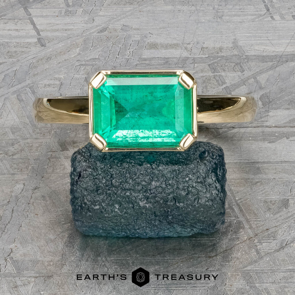 The "Marcy" Ring in 14k yellow gold features a 1.80-carat emerald-cut green gemstone resting on a rough stone surface, with "Earth's Treasury" below.