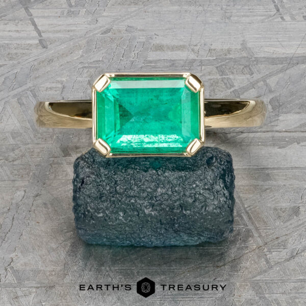 The "Marcy" Ring in 14k yellow gold with 1.80-Carat Emerald The "Marcy" Ring in 14k yellow gold features a 1.80-carat emerald-cut green gemstone resting on a rough stone surface, with "Earth's Treasury" below.