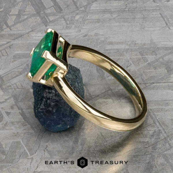 The "Marcy" Ring in 14k yellow gold with 1.80-Carat Emerald The "Marcy" Ring in 14k yellow gold with a 1.80-carat emerald stands upright, propped by a rough blue stone on a textured gray surface. "EARTH'S TREASURY" is displayed at the bottom.