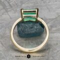 The "Marcy" Ring in 14k yellow gold with a 1.80-carat emerald stands upright before a rough blue-green stone on textured gray. "Earth's Treasury" is printed below.