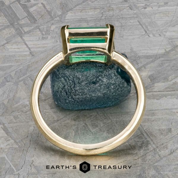 The "Marcy" Ring in 14k yellow gold with 1.80-Carat Emerald The "Marcy" Ring in 14k yellow gold with a 1.80-carat emerald stands upright before a rough blue-green stone on textured gray. "Earth's Treasury" is printed below.