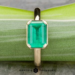 The "Marcy" Ring in 14k yellow gold showcases a 1.80-carat emerald in an upright, rectangular cut, displayed against a light green background.