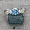 The "Lobelia" ring in platinum, featuring a 1.19-carat cushion-cut Montana sapphire and oval diamonds, is displayed on a rough blue stone surface with the text “EARTH'S TREASURY” at the bottom.
