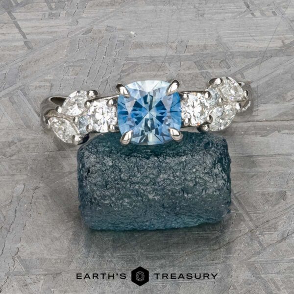 The "Lobelia" ring in platinum with 1.19-carat Montana sapphire The "Lobelia" ring in platinum, featuring a 1.19-carat cushion-cut Montana sapphire and oval diamonds, is displayed on a rough blue stone surface with the text “EARTH'S TREASURY” at the bottom.