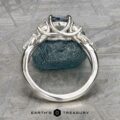 A close-up side view of the "Lobelia" ring in platinum with a 1.19-carat Montana sapphire, displayed upright against a rough blue-green stone, with the Earth's Treasury logo at the bottom.