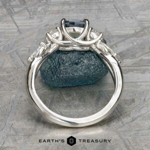 A close-up side view of the "Lobelia" ring in platinum with a 1.19-carat Montana sapphire, displayed upright against a rough blue-green stone, with the Earth's Treasury logo at the bottom.