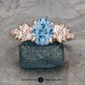 The "Celeste" ring in 14k rose gold features a 2.37-carat oval Montana sapphire and clusters of round white stones on each side, elegantly displayed on a rough blue crystal.