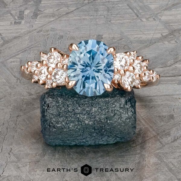 The "Celeste" ring in 14k rose gold with 2.37-carat Montana sapphire The "Celeste" ring in 14k rose gold features a 2.37-carat oval Montana sapphire and clusters of round white stones on each side, elegantly displayed on a rough blue crystal.