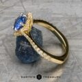 The "Plumeria" ring in 18k yellow gold and platinum with a 1.18-carat Ceylon sapphire is displayed against a rough blue crystal on a textured gray surface. Text at the bottom reads "Earth's Treasury.