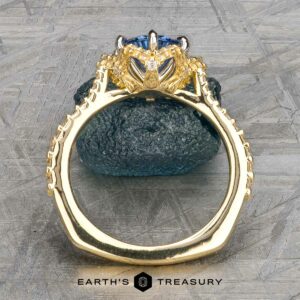 The "Plumeria" ring in 18k yellow and platinum, featuring a 1.18-carat Ceylon sapphire in an ornate prong setting, displayed upright on dark stone with “EARTH'S TREASURY” below.