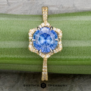 The "Plumeria" ring in 18k yellow and platinum, featuring a 1.18-carat Ceylon sapphire surrounded by diamonds, is displayed on a green plant stem with the "Earth's Treasury" logo below.