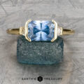 The "Dinara" ring in 18k yellow gold, featuring a 2.20-carat rectangular Montana sapphire and two smaller clear stones, rests on a textured green surface.