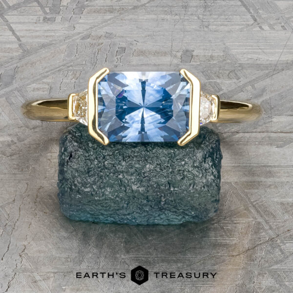 The "Dinara" ring in 18k yellow gold with 2.20-carat Montana sapphire The "Dinara" ring in 18k yellow gold, featuring a 2.20-carat rectangular Montana sapphire and two smaller clear stones, rests on a textured green surface.