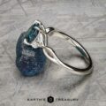 The "Anise" ring in platinum, featuring a 1.37-carat Montana sapphire and two clear side stones, is shown resting on a rough blue mineral against a textured backdrop.
