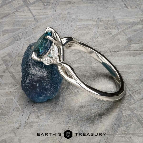 The "Anise" ring in platinum with 1.37-Carat Montana Sapphire The "Anise" ring in platinum, featuring a 1.37-carat Montana sapphire and two clear side stones, is shown resting on a rough blue mineral against a textured backdrop.