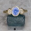 The "Kaelin" Ring in 14k yellow gold features a 1.00-carat Ceylon sapphire center, flanked by two oval clear gems, shown on a rough dark stone with Earth's Treasury branding below.