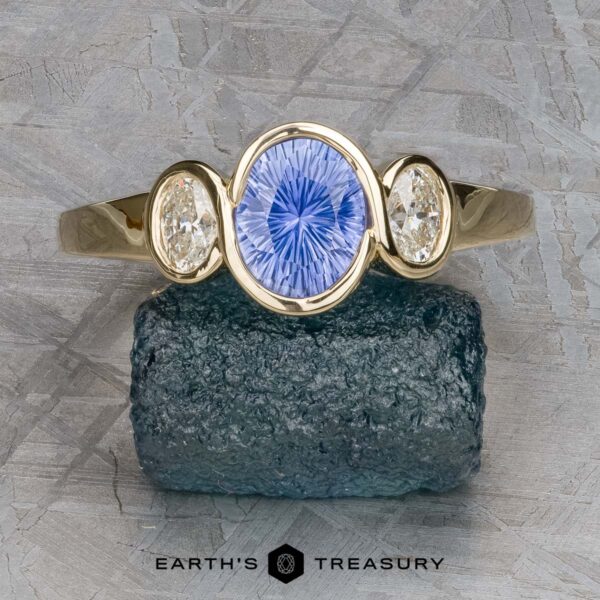 The "Kaelin" Ring in 14k yellow gold with 1.00-carat Ceylon sapphire The "Kaelin" Ring in 14k yellow gold features a 1.00-carat Ceylon sapphire center, flanked by two oval clear gems, shown on a rough dark stone with Earth's Treasury branding below.