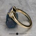 The "Kaelin" Ring in 14k yellow gold with a 1.00-carat Ceylon sapphire is displayed propped on a rough dark blue stone against a textured gray background. "EARTH'S TREASURY" appears at the bottom.