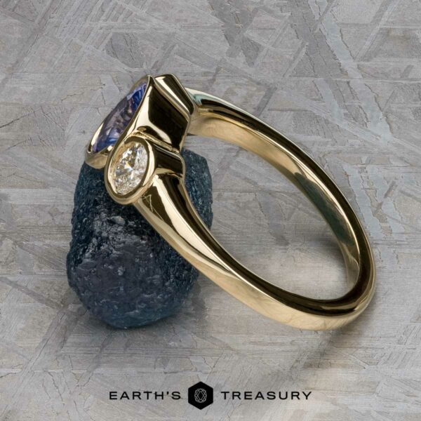 The "Kaelin" Ring in 14k yellow gold with 1.00-carat Ceylon sapphire The "Kaelin" Ring in 14k yellow gold with a 1.00-carat Ceylon sapphire is displayed propped on a rough dark blue stone against a textured gray background. "EARTH'S TREASURY" appears at the bottom.