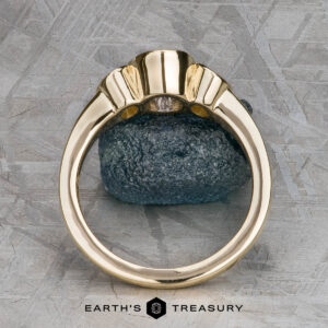 The "Kaelin" Ring in 14k yellow gold with a 1.00-carat Ceylon sapphire is shown upright in a prong setting on a dark stone background, viewed from the side. The "Earth's Treasury" logo appears at the bottom.