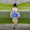 The "Kaelin" Ring in 14k yellow gold features a 1.00-carat round Ceylon sapphire center, flanked by two oval white stones, displayed on a green leaf background.