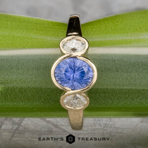 The "Kaelin" Ring in 14k yellow gold features a 1.00-carat round Ceylon sapphire center, flanked by two oval white stones, displayed on a green leaf background.