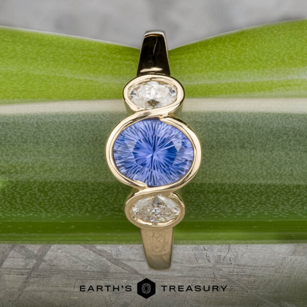 The "Kaelin" Ring in 14k yellow gold with 1.00-carat Ceylon sapphire The "Kaelin" Ring in 14k yellow gold features a 1.00-carat round Ceylon sapphire center, flanked by two oval white stones, displayed on a green leaf background.