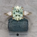 The "Dryad" ring in 14k yellow gold, featuring a 3.50-carat oval light green Montana sapphire and two side diamonds, is displayed on rough dark stone with a textured gray background and "EARTH'S TREASURY" text at the bottom.