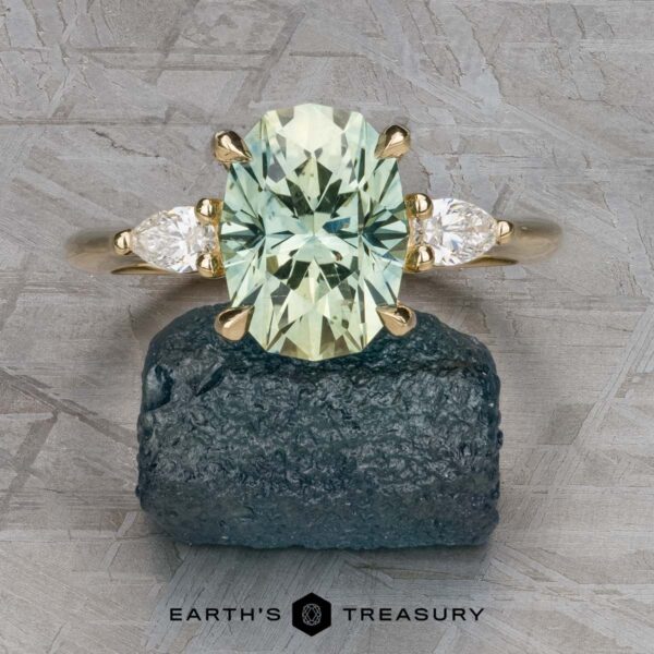 The "Dryad" ring in 14k yellow gold with 3.50-carat Montana sapphire The "Dryad" ring in 14k yellow gold, featuring a 3.50-carat oval light green Montana sapphire and two side diamonds, is displayed on rough dark stone with a textured gray background and "EARTH'S TREASURY" text at the bottom.