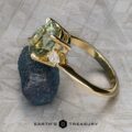The "Dryad" ring in 14k yellow gold, featuring a 3.50-carat light green Montana sapphire and a small white diamond, rests on a rough, dark stone surface. The Earth's Treasury logo appears at the bottom.