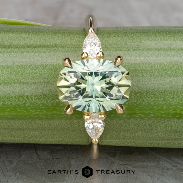 The "Dryad" ring in 14k yellow gold with 3.50-carat Montana sapphire The "Dryad" ring in 14k yellow gold features a 3.50-carat oval Montana sapphire and two pear-shaped clear stones, shown on a green stem with the "Earth's Treasury" logo below.