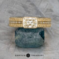 The "Augusta" ring in 14k yellow gold has a 1.01-carat emerald-cut diamond, baguette side stones, and intricate details, elegantly displayed upright on a green mineral stone.