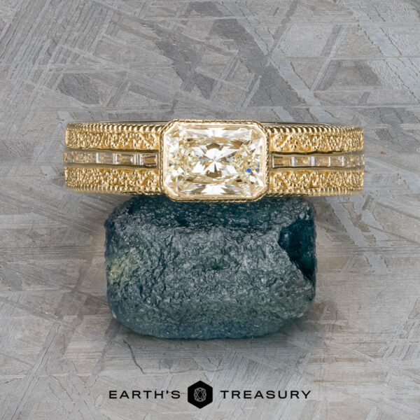 The "Augusta" ring in 14k yellow gold with 1.01-carat diamond The "Augusta" ring in 14k yellow gold has a 1.01-carat emerald-cut diamond, baguette side stones, and intricate details, elegantly displayed upright on a green mineral stone.