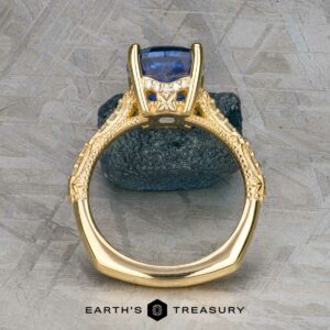 The "Marilyn" ring in 18k yellow gold features an intricate band and a 3.20-carat Ceylon sapphire in a four-prong setting, shown upright on a textured gray surface.