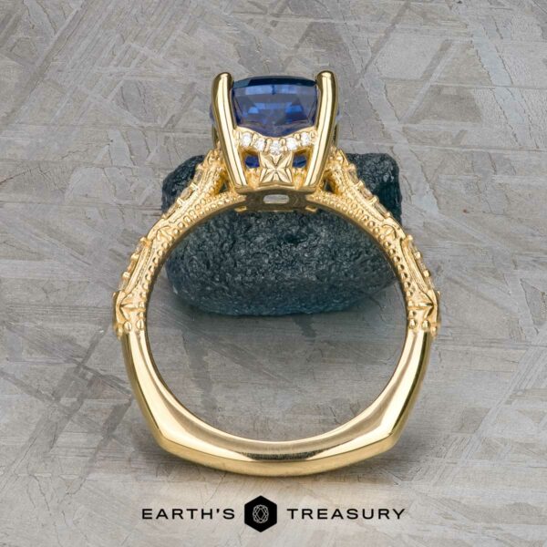 The "Marilyn" ring in 18k yellow gold with 3.20-carat Ceylon sapphire The "Marilyn" ring in 18k yellow gold features an intricate band and a 3.20-carat Ceylon sapphire in a four-prong setting, shown upright on a textured gray surface.