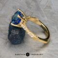 The "Marilyn" ring in 18k yellow gold, featuring a 3.20-carat Ceylon sapphire and diamond-accented band, is displayed upright on rough dark stone against a textured gray background.