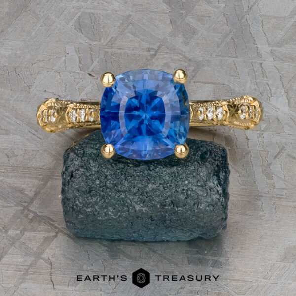 The "Marilyn" ring in 18k yellow gold with 3.20-carat Ceylon sapphire The "Marilyn" ring in 18k yellow gold, featuring a 3.20-carat Ceylon sapphire and diamond accents, is displayed on a rough dark stone with the Earth's Treasury logo below.