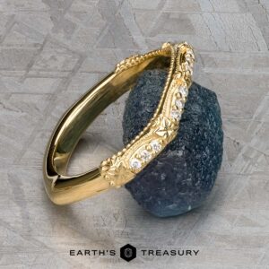 The "Marilyn" band in 18k yellow gold, featuring diamond accents and floral details, is shown encircling a rough blue gemstone on a gray textured surface.