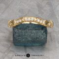 The "Marilyn" band in 18k yellow gold, featuring diamond accents and ornate detailing, is showcased on a dark textured mineral surface with a geometric metallic background.