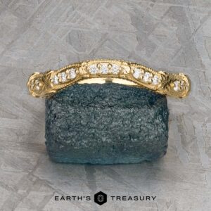 The "Marilyn" band in 18k yellow gold, featuring diamond accents and ornate detailing, is showcased on a dark textured mineral surface with a geometric metallic background.