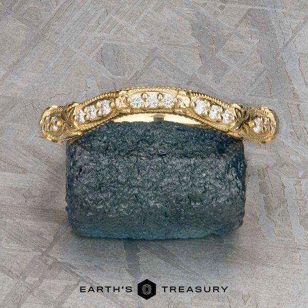 The "Marilyn" band in 18k yellow gold The "Marilyn" band in 18k yellow gold, featuring diamond accents and ornate detailing, is showcased on a dark textured mineral surface with a geometric metallic background.