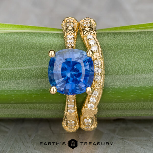 The "Marilyn" ring in 18k yellow gold with 3.20-carat Ceylon sapphire, alongside the "Marilyn" band in 18k yellow gold The "Marilyn" ring in 18k yellow gold with a 3.20-carat Ceylon sapphire is paired with the "Marilyn" band in 18k yellow gold, both displayed on a green stem with Earth's Treasury branding below.