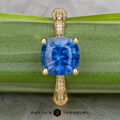 The "Marilyn" ring in 18k yellow gold features a 3.20-carat cushion-cut Ceylon sapphire and diamond-accented band, displayed on a green leaf with Earth’s Treasury logo at the bottom.