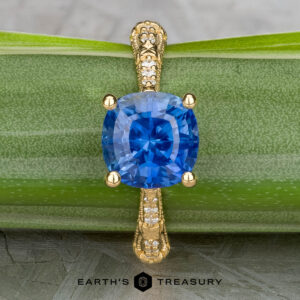 The "Marilyn" ring in 18k yellow gold features a 3.20-carat cushion-cut Ceylon sapphire and diamond-accented band, displayed on a green leaf with Earth’s Treasury logo at the bottom.