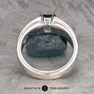 The "Shannon" ring in platinum with a 1.06-carat Montana Sapphire is displayed upright, highlighting the blue rectangular stone and band against a textured metallic background. The "Earth's Treasury" logo appears below.