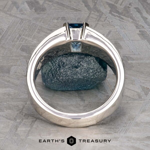 The "Shannon" ring in platinum with 1.06-Carat Montana Sapphire The "Shannon" ring in platinum with a 1.06-carat Montana Sapphire is displayed upright, highlighting the blue rectangular stone and band against a textured metallic background. The "Earth's Treasury" logo appears below.