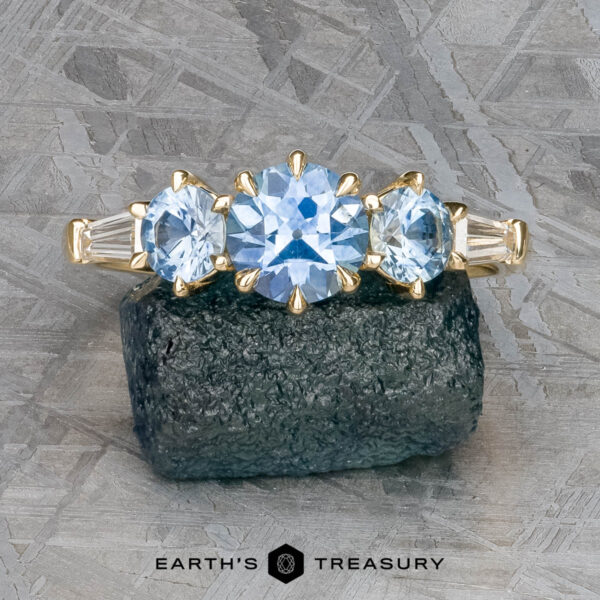 The "Concordia" ring in 14k yellow gold with 0.94-carat Montana sapphire The "Concordia" ring in 14k yellow gold, featuring a 0.94-carat Montana sapphire and baguette side stones, is displayed on a rough, dark green mineral with a textured gray background.