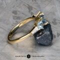 The "Concordia" ring in 14k yellow gold, featuring a 0.94-carat Montana sapphire and baguette diamonds, is displayed against a rough mineral sample on a textured surface. The Earth's Treasury logo appears at the bottom.