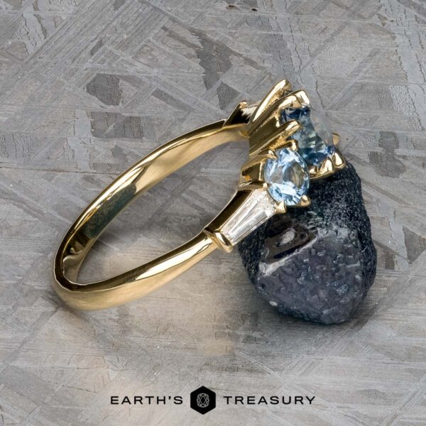 The "Concordia" ring in 14k yellow gold with 0.94-carat Montana sapphire The "Concordia" ring in 14k yellow gold, featuring a 0.94-carat Montana sapphire and baguette diamonds, is displayed against a rough mineral sample on a textured surface. The Earth's Treasury logo appears at the bottom.